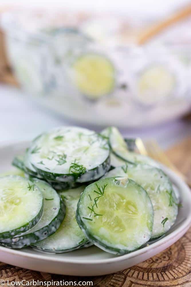 close up of cucumber salad on a white plate with a fork on the side and more cucumber salad in a clear bowl in the background