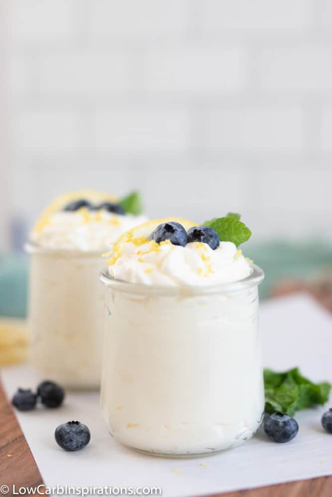 close up of keto lemon mousse in a small clear glass with a lemon slice, blueberries and mint on a table