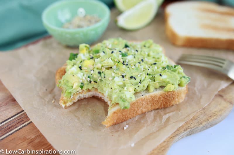 low carb bread topped with avocado egg salad on a wood cutting board with lies and seasonings in the background with a bite taken out of the sandwich