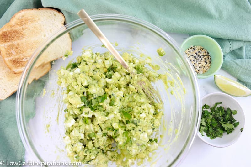 mixed avocado egg salad recipe in a clear bowl with extra seasonings and low carb bread on the side