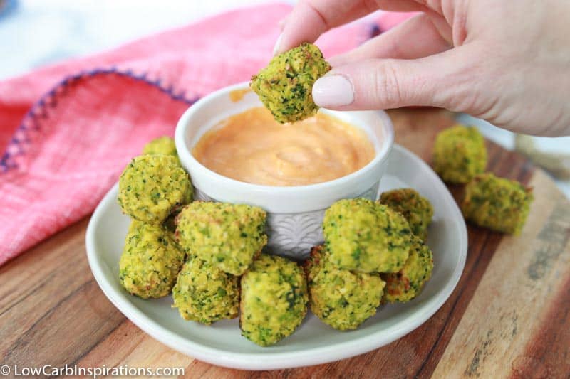 baked broccoli tots recipe a white plate with a white bowl of dipping sauce and a handing holding one close up