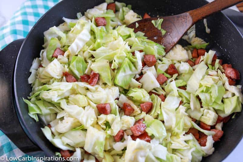 adding cabbage and cooking sausage and cabbage with a wooden spoon in a skillet