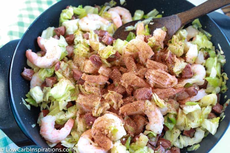 adding seasoning and cooking sausage, shrimp and cabbage cooking in a skillet with a wooden spoon