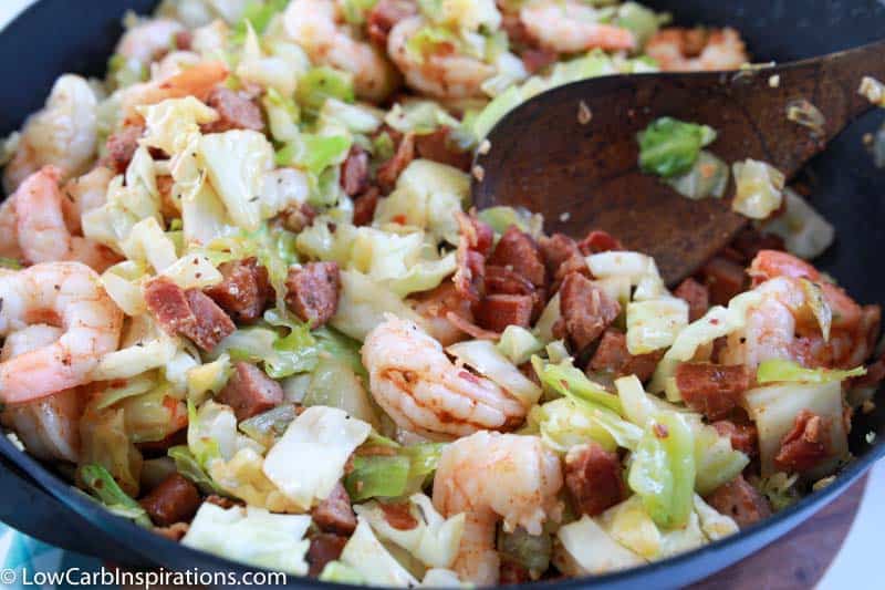 cooking sausage, shrimp and cabbage cooking in a skillet with a wooden spoon