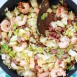 overhead photo of sausage, shrimp and cabbage cooking in a skillet with a wooden spoon