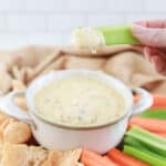 close up of keto cheese dip on a celery stick with white cheese sauce with carrots, celery and pork rinds in the background