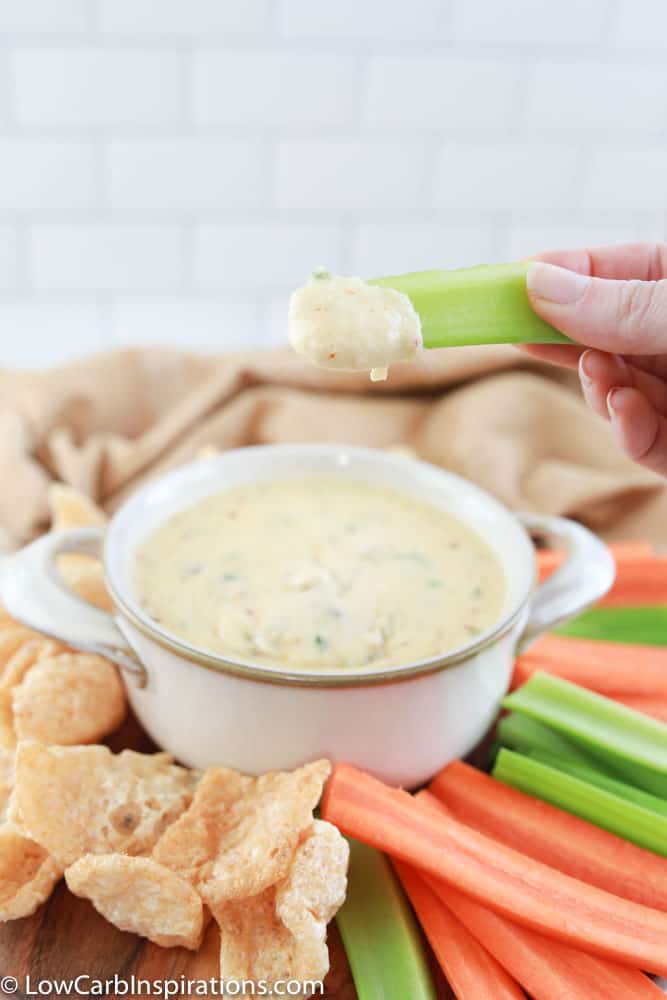 close up of keto cheese dip on a celery stick with white cheese sauce with carrots, celery and pork rinds in the background