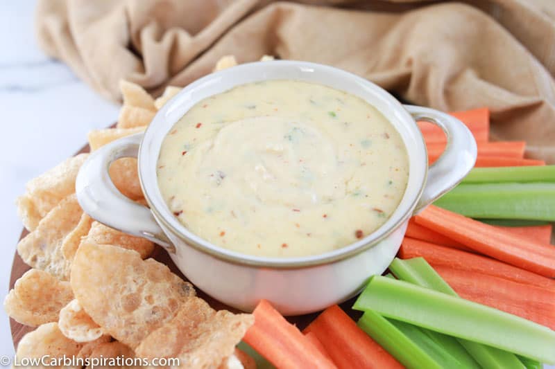keto queso dip in a white bowl surrounded with celery, carrots, and pork rinds on a table with a tan towel in the background