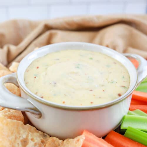 close up of keto queso dip in a white bowl surrounded with celery, carrots, and pork rinds on a table with a tan towel in the background