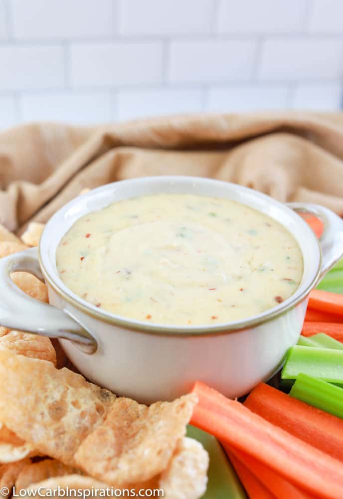 close up of keto queso dip in a white bowl surrounded with celery, carrots, and pork rinds on a table with a tan towel in the background