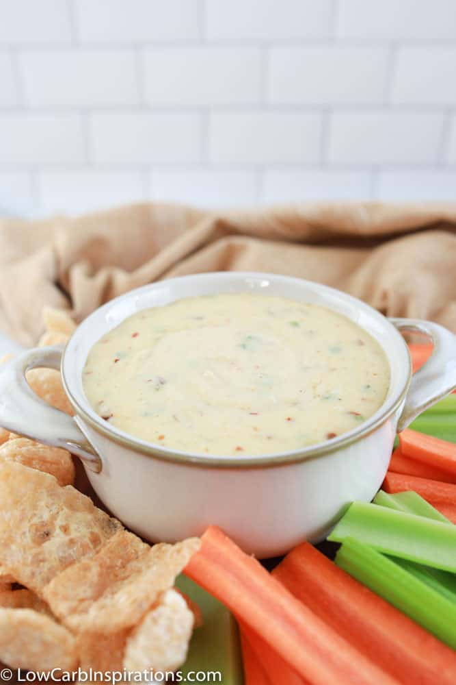 keto cheese dip in a white bowl surrounded with celery, carrots, and pork rinds on a table with a tan towel in the background