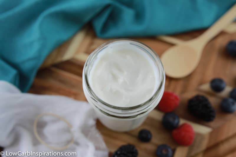 thickened coconut yogurt in a mason jar with a piece of cheesecloth, wooden spoon and mixed berries on the side