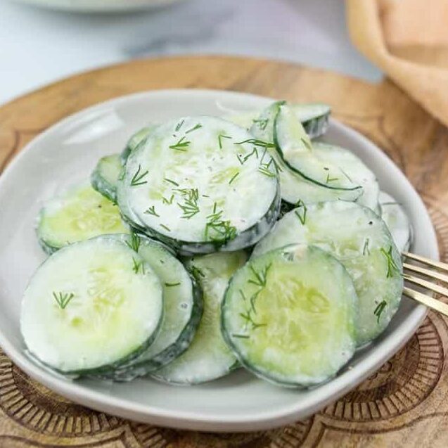 cucumber salad with dill on a white plate with a fork on the side and more cucumber salad in a clear bowl in the background