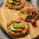 angled photo of cauliflower pancakes on a wood cutting board topped with avocado and tomatoes