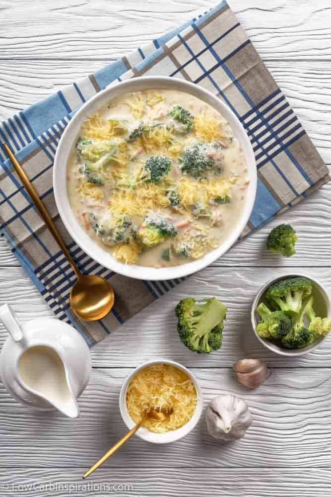 cheesy broccoli and vegetable soup in a white bowl with a golden spoon on a napkin and ingredients, vertical view from above, flat lay